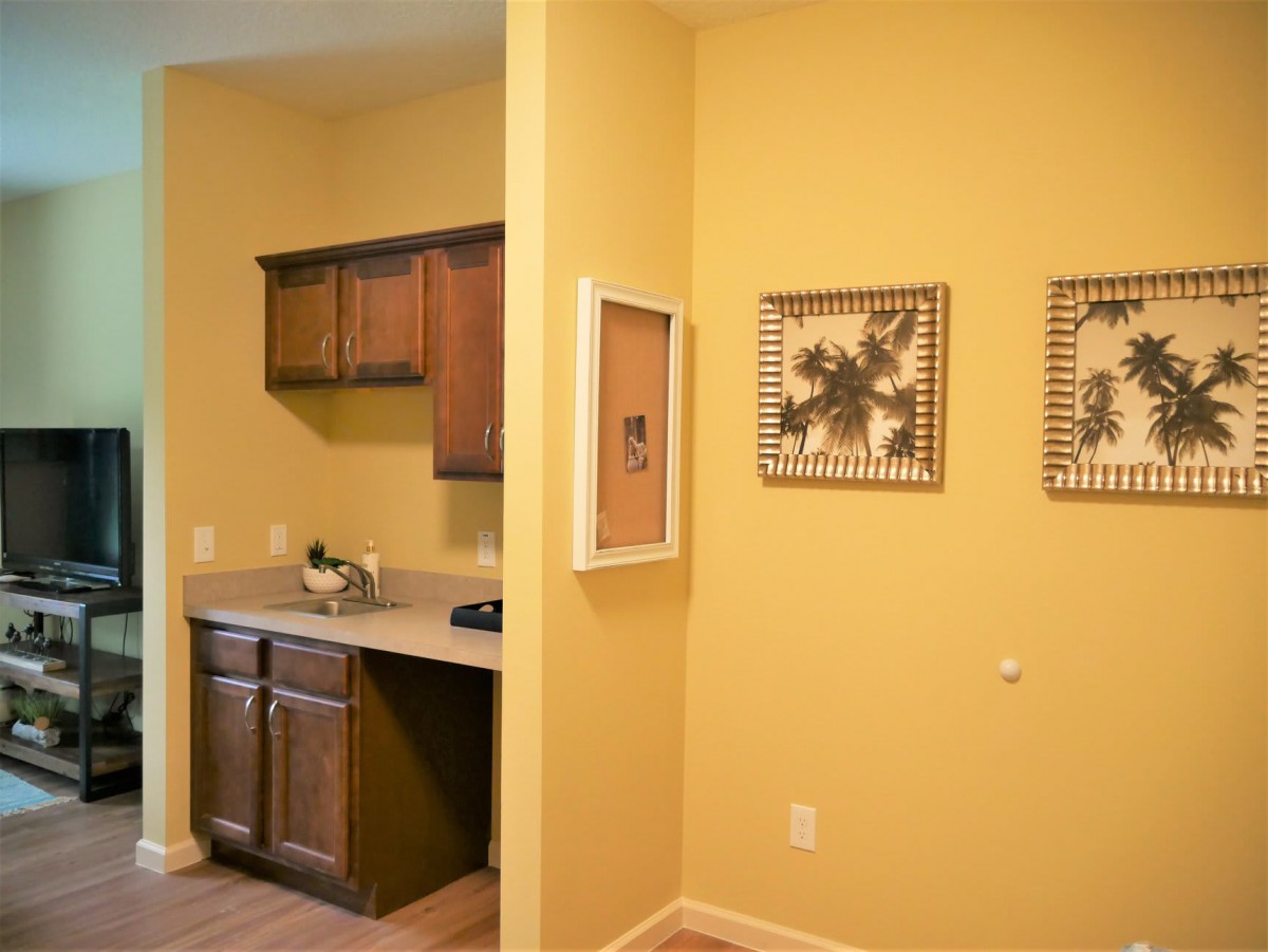 A cozy kitchen area with yellow walls and wooden cabinets, adorned with framed artwork.