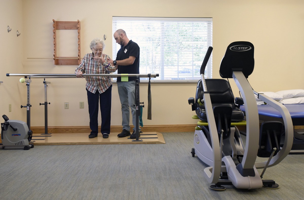 A therapist assists an elderly woman with exercises using parallel bars in a rehabilitation facility.