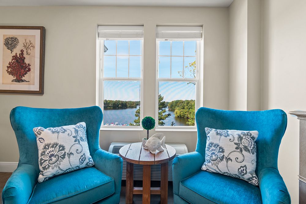 A cozy living room setup featuring two blue chairs with patterned cushions, flanking a small wooden table, overlooking a scenic view through large windows.