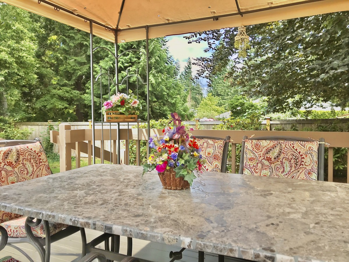 A tranquil patio setup featuring a marble table adorned with a vibrant flower arrangement, surrounded by lush greenery under a canopy.