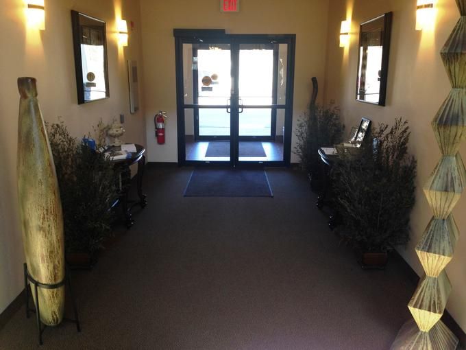 A well-lit hallway leading to a glass door entrance, adorned with plants and decorated with framed artwork on the walls.