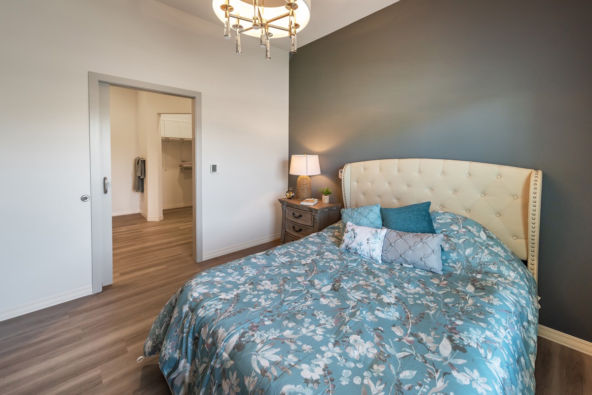 A cozy bedroom featuring a floral-patterned bed with decorative pillows, a nightstand with a lamp, and a stylish chandelier.