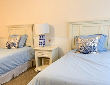 A cozy bedroom with two twin beds featuring blue bedding and decorative pillows, separated by a white nightstand with a lamp.