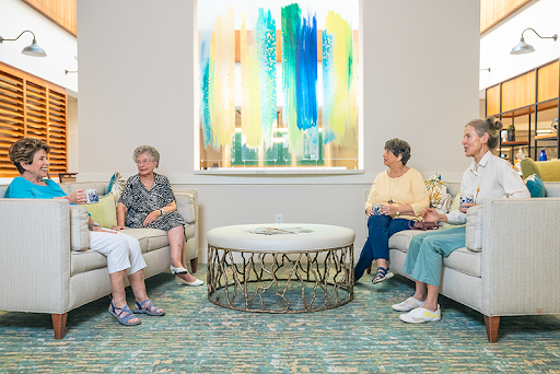 A cozy gathering of four senior women enjoying a conversation in a well-lit lounge area adorned with colorful artwork.