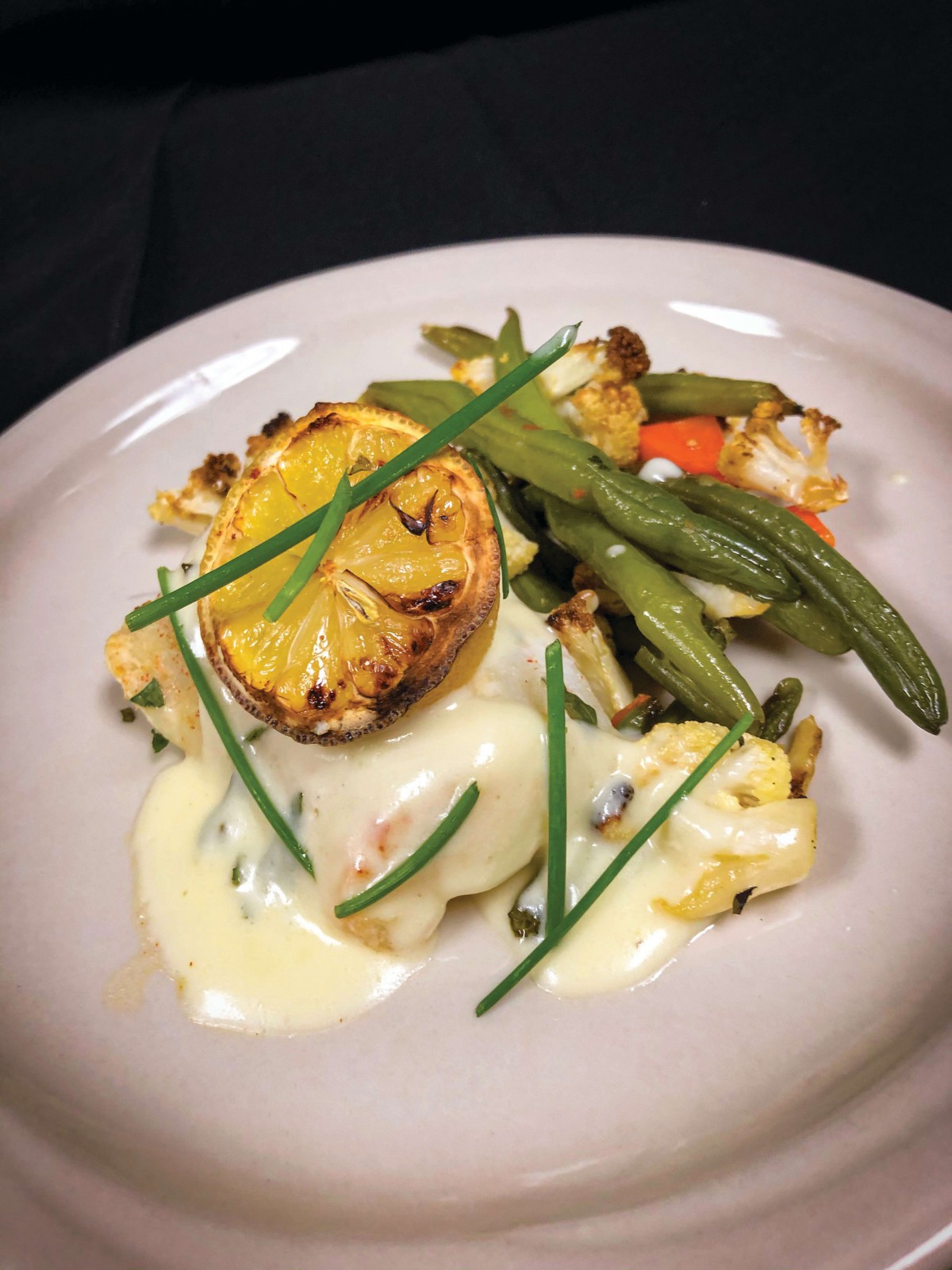 A gourmet dish featuring grilled lemon, green beans, and vegetables topped with a creamy sauce and garnished with chives.