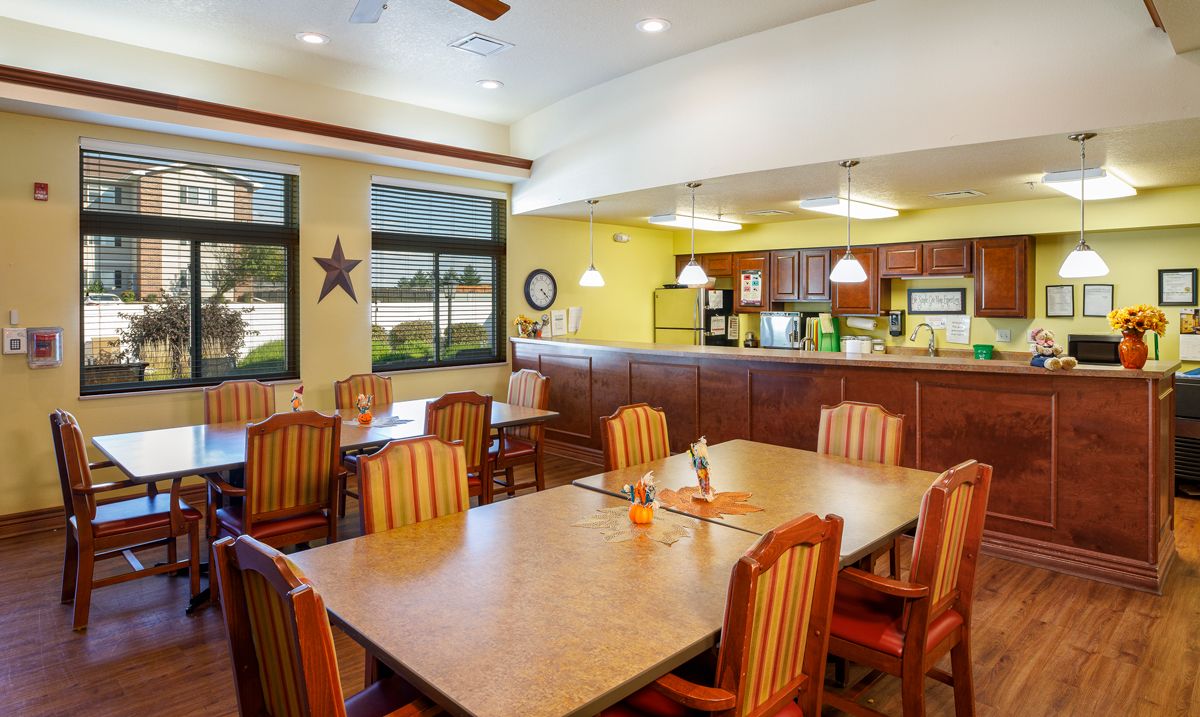 A cozy dining area with wooden chairs and tables, adjacent to a kitchen, featuring warm lighting and large windows.