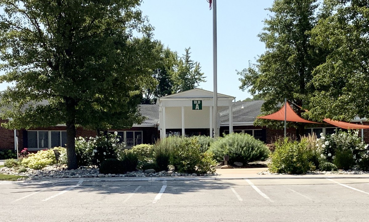 The image shows the exterior of a building surrounded by trees and landscaping, with a parking lot in front.