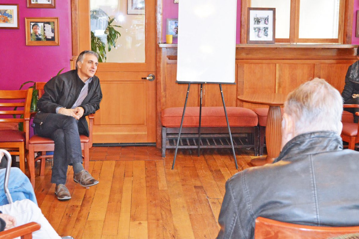 A man seated, engaging in a discussion with another person in a room equipped with a whiteboard.
