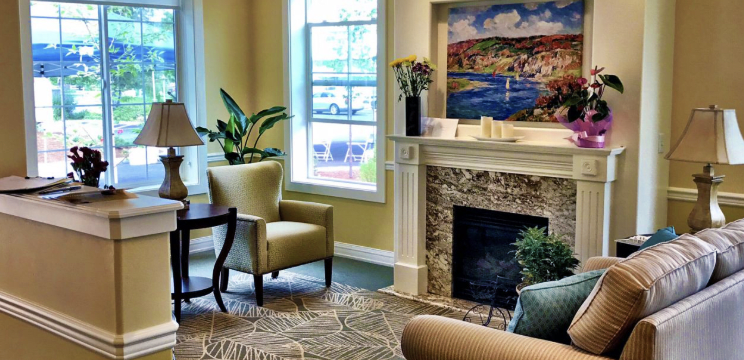 A cozy living room featuring an armchair and a fireplace, adorned with a painting and surrounded by plants.