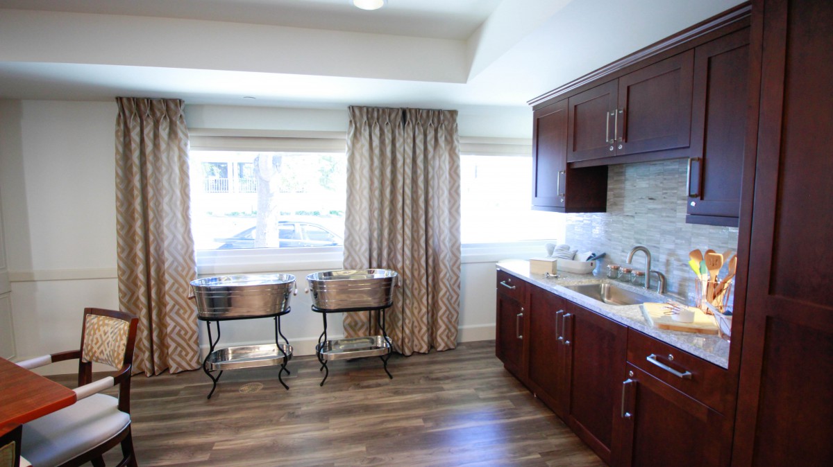 A bright kitchen space featuring wooden cabinets, large windows with curtains, and sleek flooring.