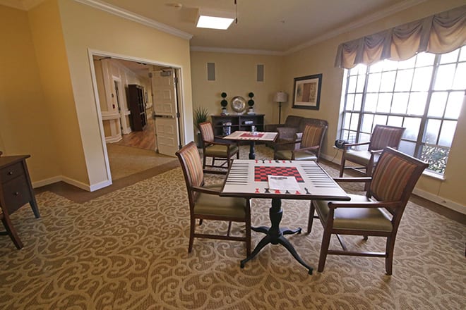 A cozy living area with tables and chairs arranged on a patterned carpet, featuring a large window allowing natural light to enter.