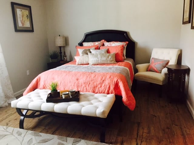 A cozy bedroom with a neatly made bed, featuring coral bedding and decorative pillows, accompanied by an armchair and a small lamp.