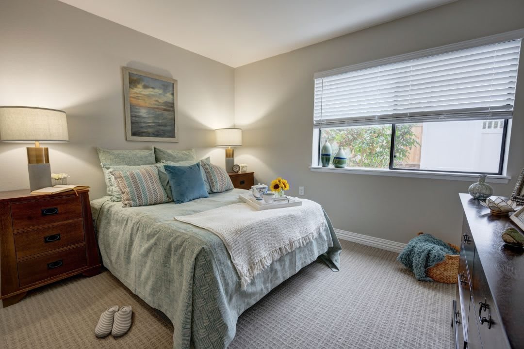 A cozy and well-lit bedroom with a neatly made bed, lamp, and window bringing in natural light.