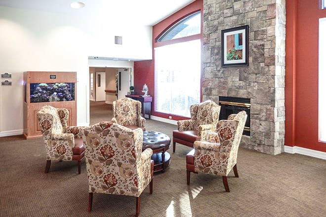 A cozy living room featuring patterned armchairs around a table, a stone fireplace, and a large window.