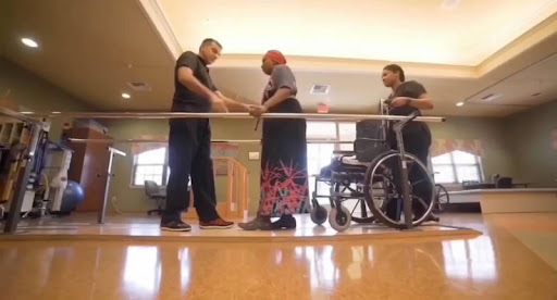 A healthcare professional assists a patient in rehabilitation, while another caregiver stands by with a wheelchair.