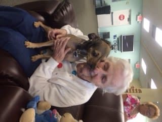 An elderly person smiling while cuddling a small dog indoors.