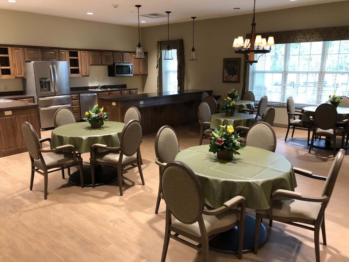 A cozy dining area with tables and chairs, featuring a kitchen in the background and decorative plants on the tables.
