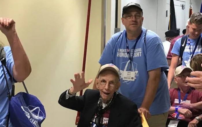 An elderly man wearing a baseball cap waves while another man stands behind him indoors.