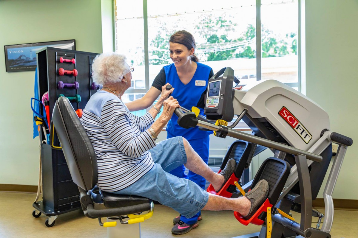 A healthcare professional assists an elderly woman with exercise using specialized equipment in a physical therapy setting.