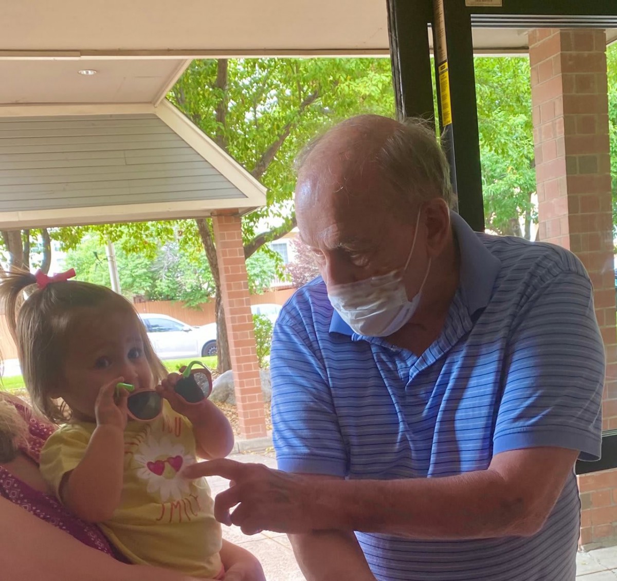 An elderly man wearing a mask interacts with a baby indoors.