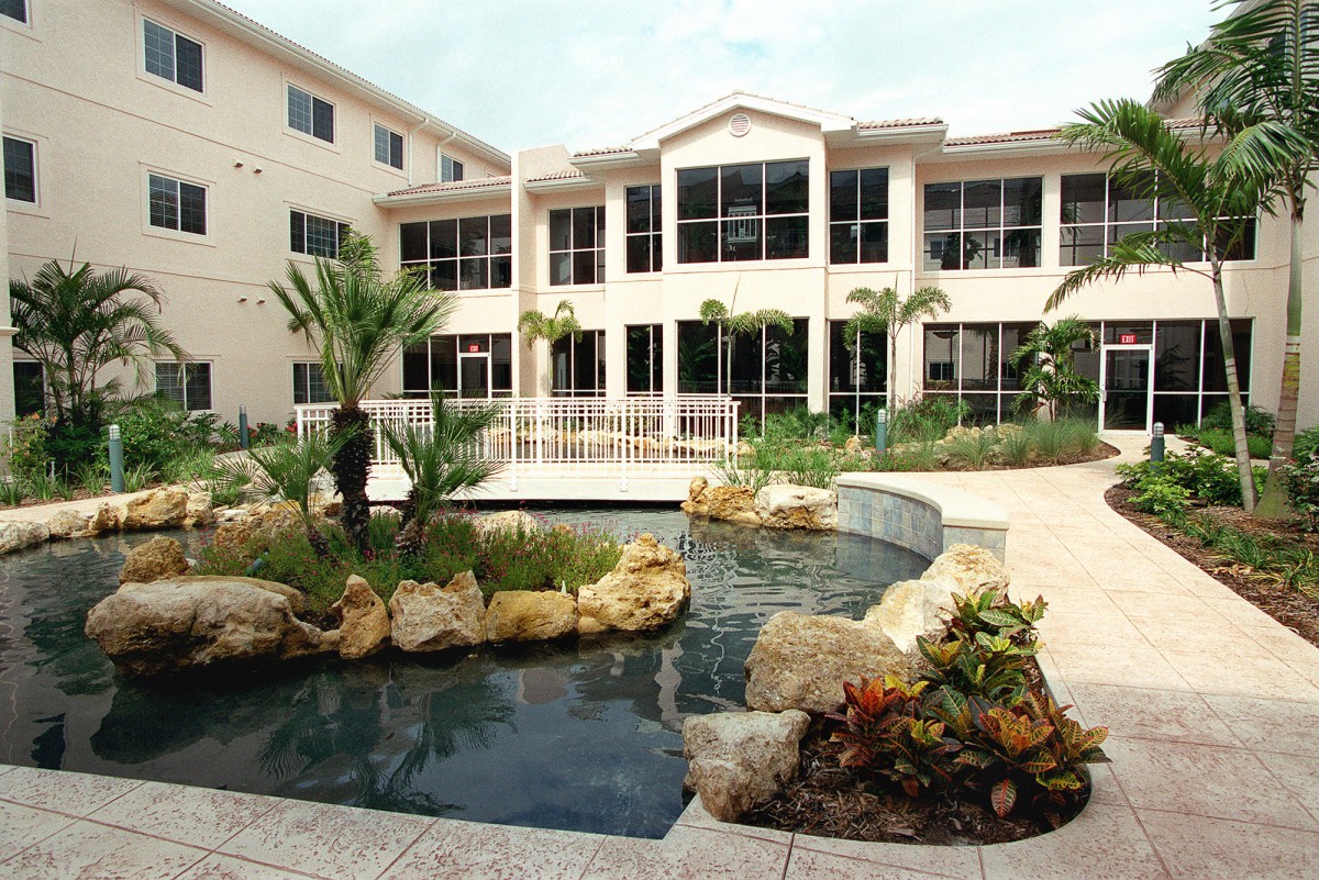 A residential building with a landscaped garden and pond in the foreground, featuring palm trees and rocks.