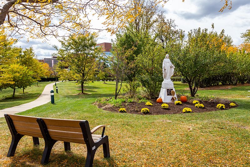 A peaceful park scene with a bench facing a statue surrounded by trees and flowers during autumn.