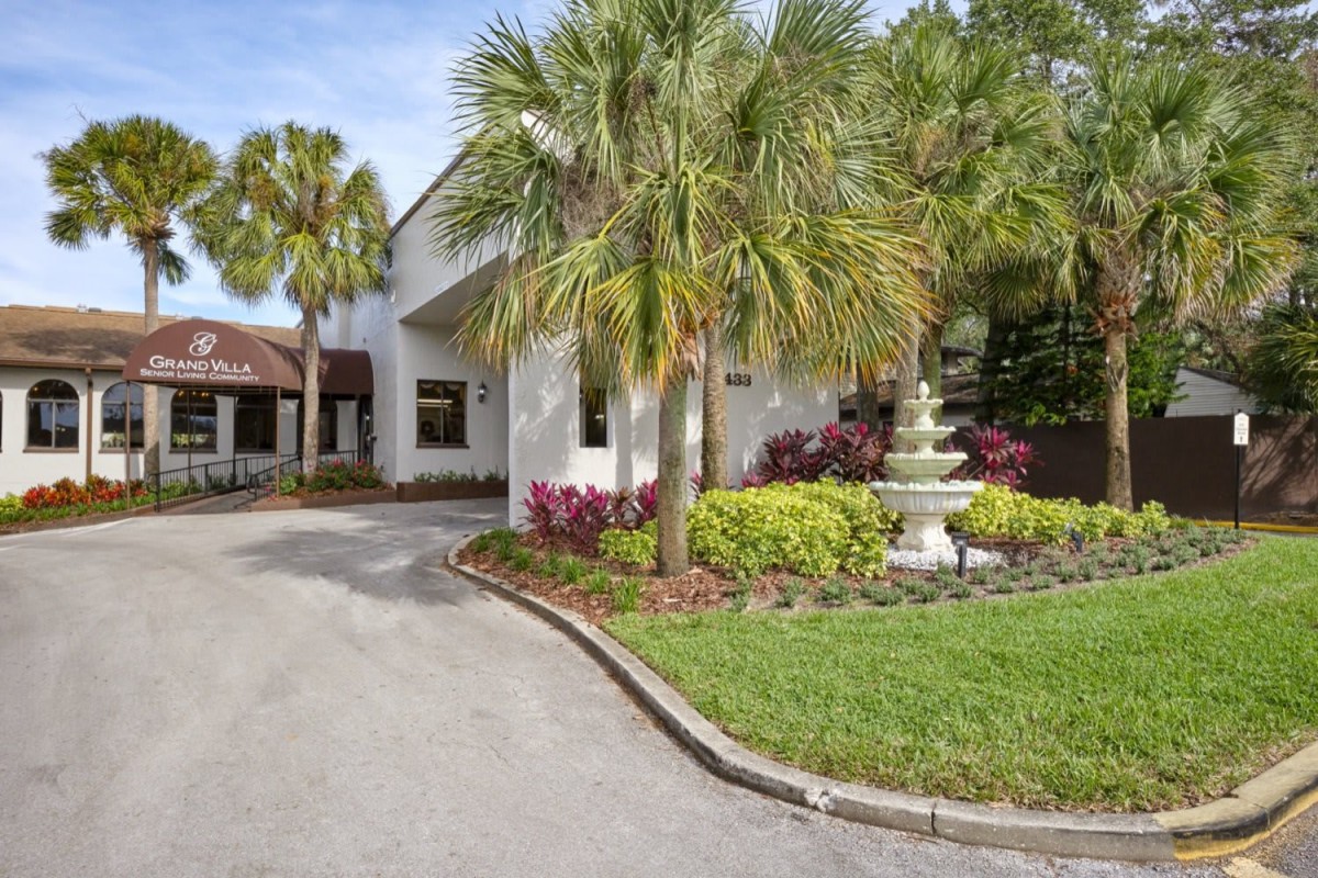 A house with a circular driveway, surrounded by palm trees and a lush garden featuring a fountain.