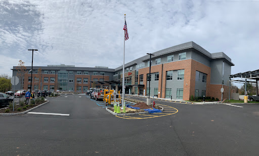 A modern hospital building featuring a flagpole in the parking area, set against a cloudy sky.