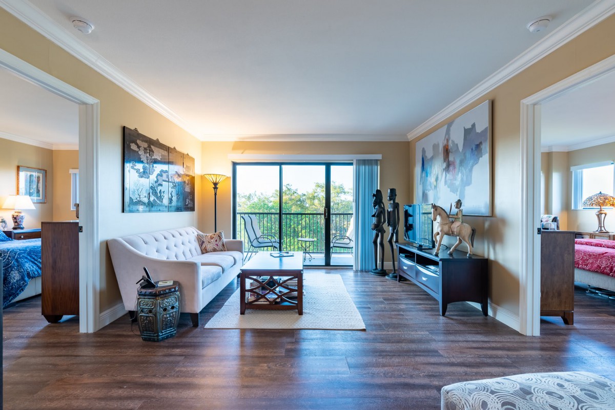 A cozy living room with modern decor, featuring a comfortable sofa, a television, and large artwork, illuminated by natural light from the balcony doors.