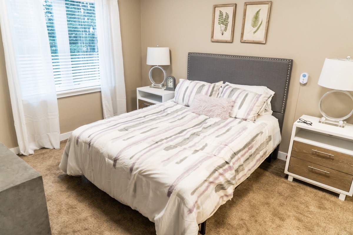 A cozy bedroom with a neatly made bed, featuring striped bedding and a gray headboard. The room includes nightstands with lamps and framed artwork above the bed.