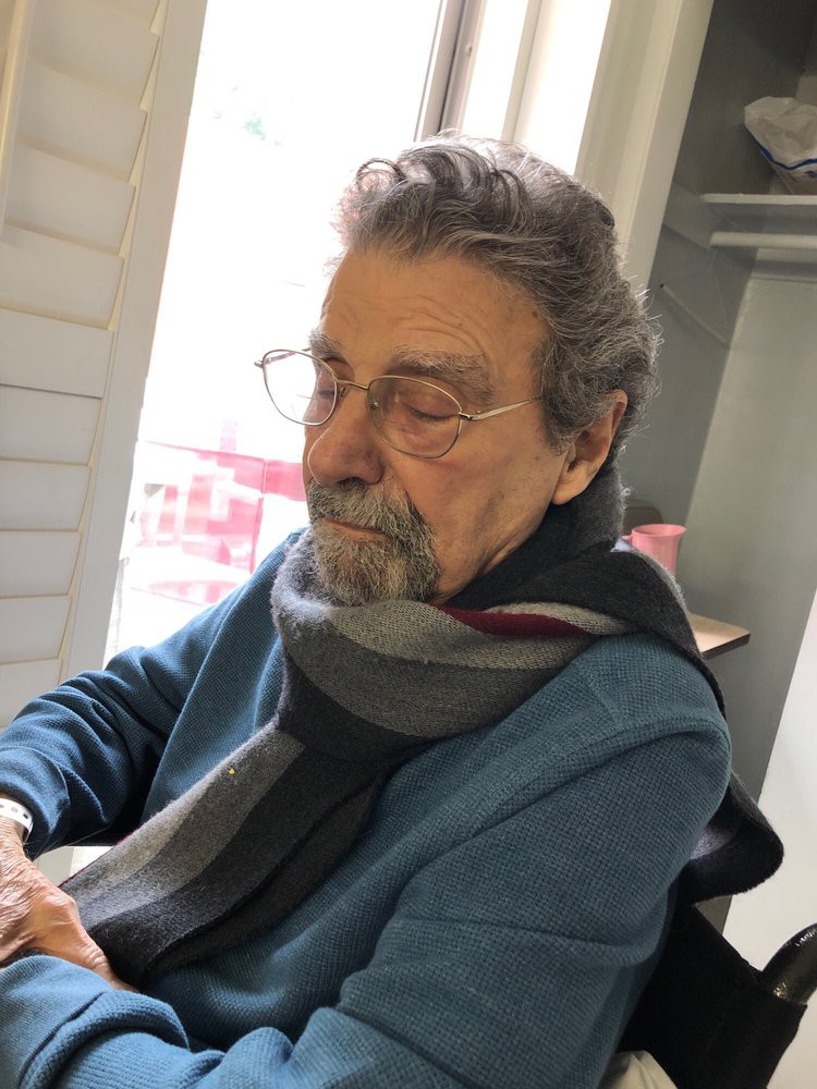 An elderly man wearing a scarf and glasses sits indoors, appearing thoughtful.