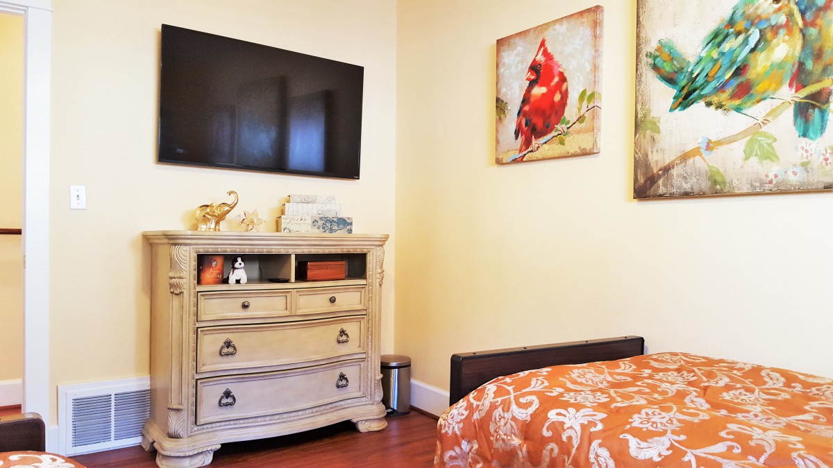 A cozy bedroom featuring a television mounted above a dresser, with colorful artwork and vibrant orange bedding.