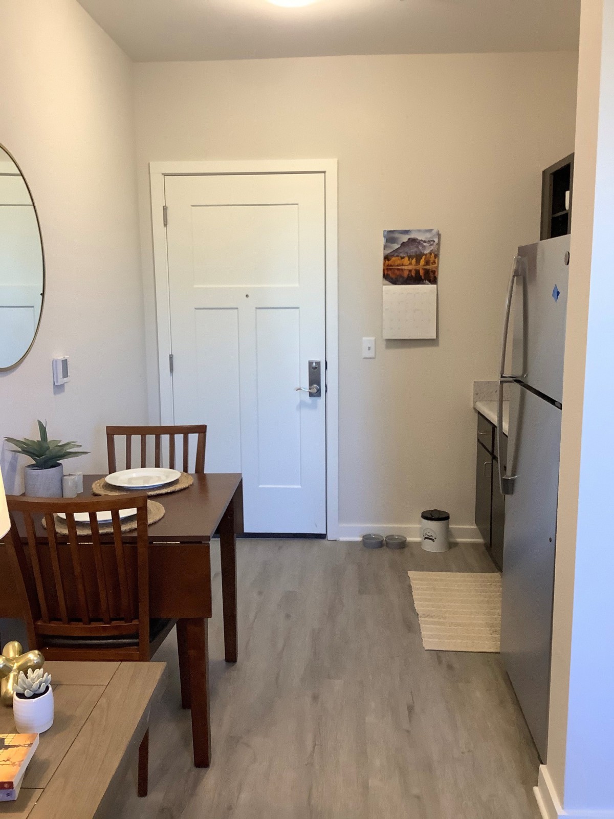 A cozy apartment kitchen with a small dining table and chairs, featuring a refrigerator and a round mirror on the wall.