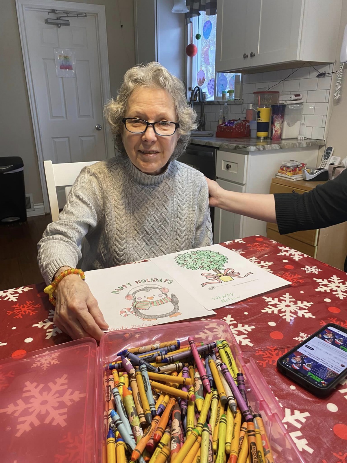 An elderly woman is sitting at a table, smiling as she shows her coloring book. A box of crayons is on the table.