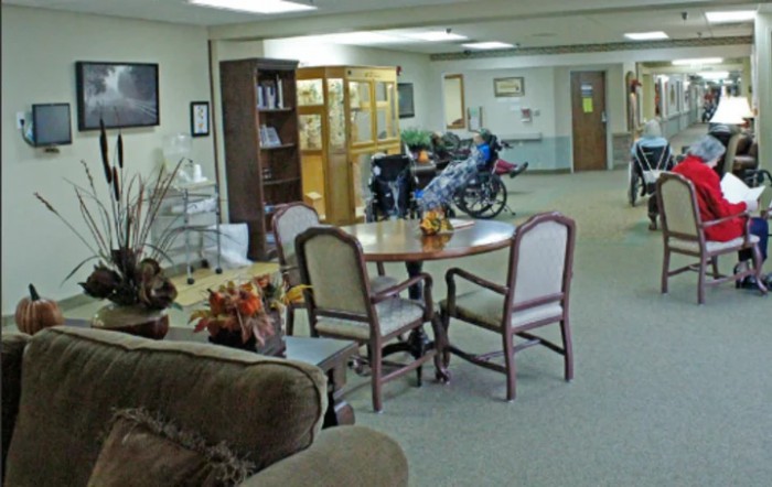 A common area in a nursing home with a table, chairs, and a person in a wheelchair.
