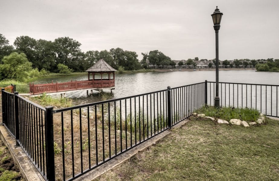 A tranquil lakeside view with a gazebo, fence, and lamp post under a cloudy sky.