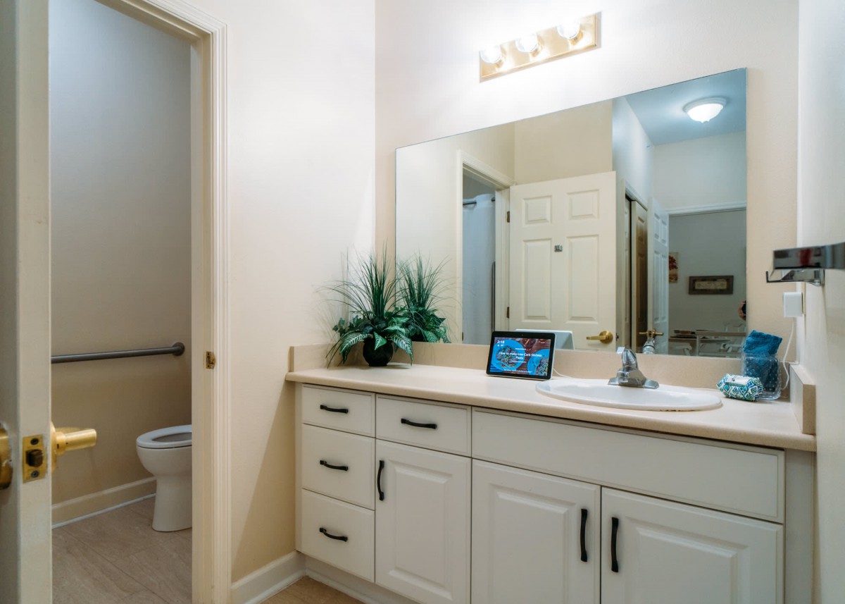 A modern bathroom with a large mirror, white cabinets, and a tablet on the counter.