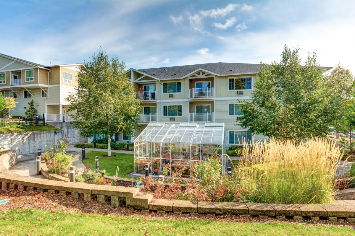 A beautiful apartment complex with a greenhouse and garden in the foreground, set against a clear blue sky.