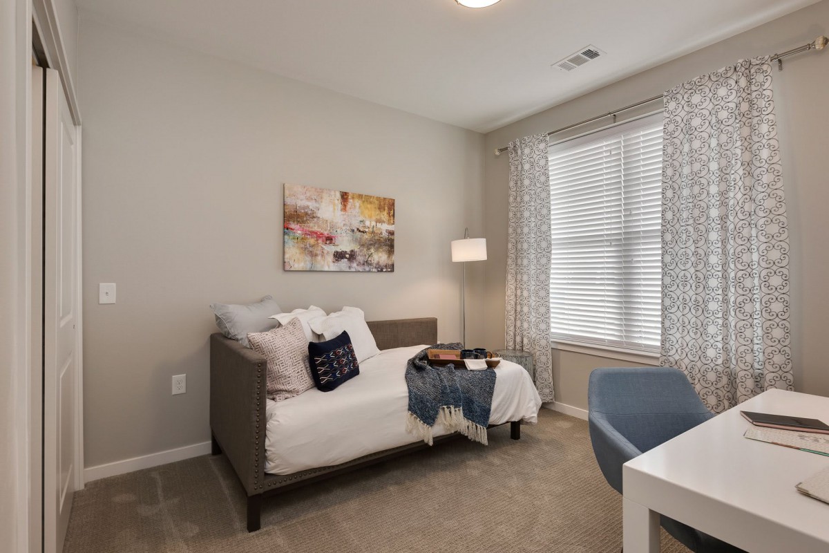 A modern bedroom featuring a daybed with decorative pillows, illuminated by natural light from large windows with blinds.