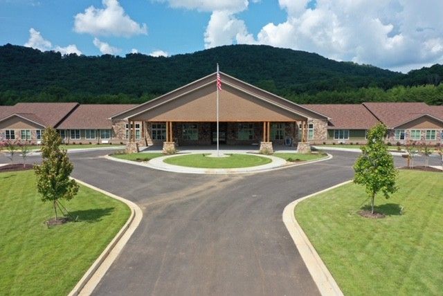 A scenic view of a large building with a driveway, set against a backdrop of mountains and a partly cloudy sky.