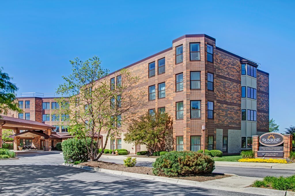 A modern brick building with large windows, surrounded by trees and neatly landscaped greenery.