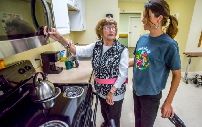 An elderly woman receives help from a caregiver in the kitchen.