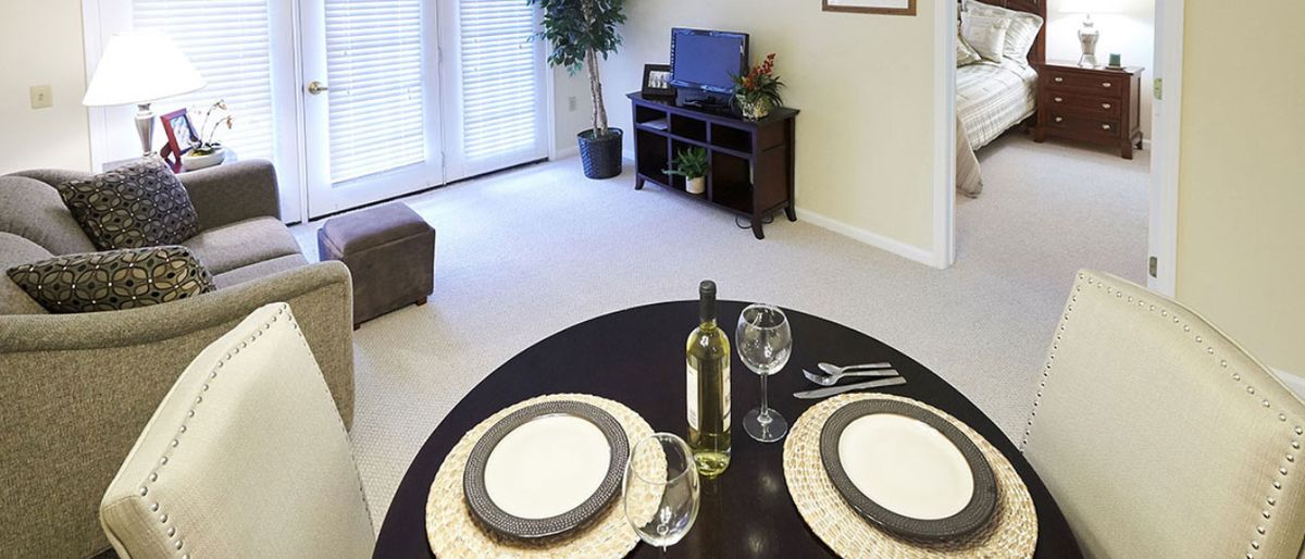 A cozy dining area set with a table for two, featuring wine glasses and a bottle, adjacent to a modern living room.
