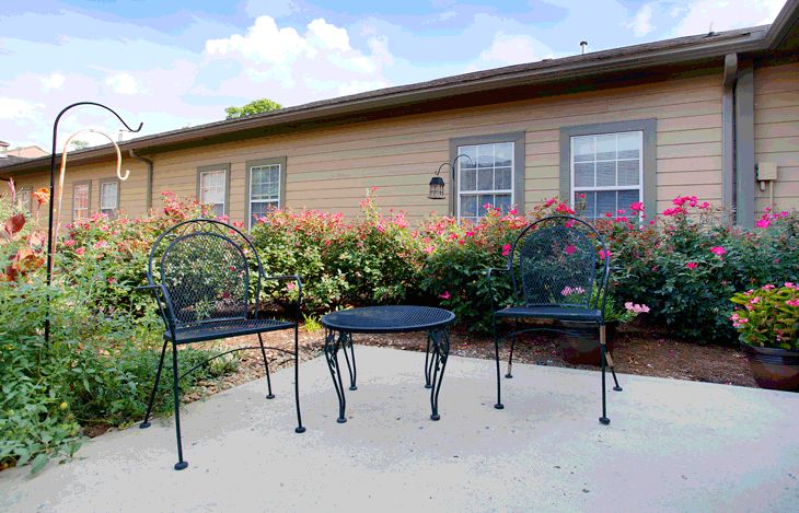 An outdoor patio area with metal chairs and a table in front of a house, surrounded by bushes.