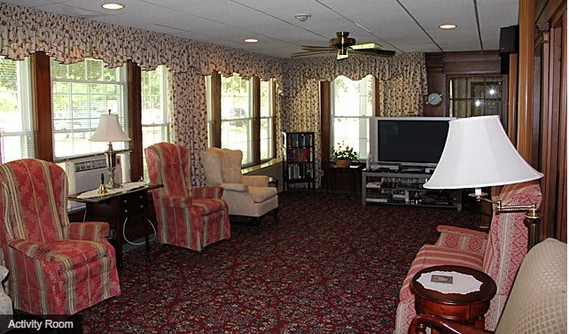 A cozy living room with patterned wallpaper and carpet, featuring multiple chairs, a television, and large windows.