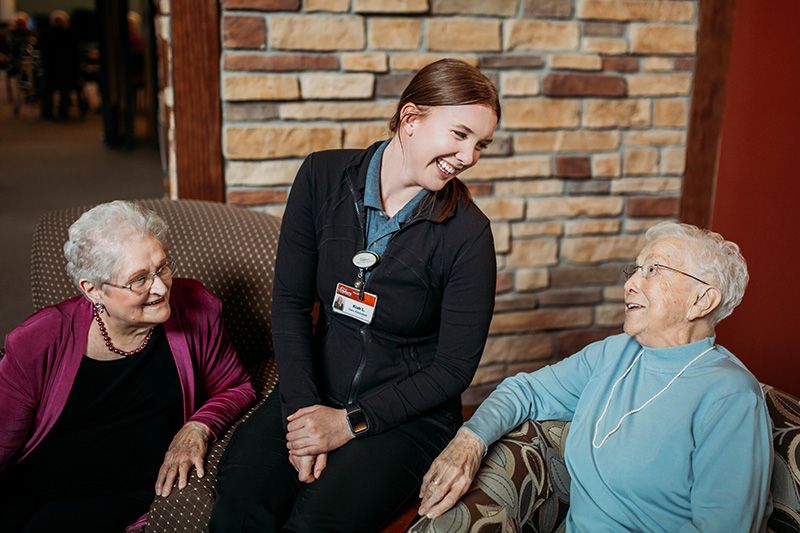 A caregiver smiling and interacting with two elderly women in a cozy setting.