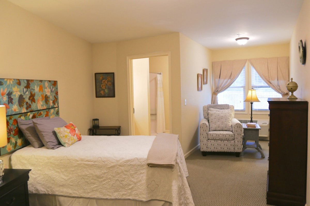 A cozy bedroom interior with a bed, chair, and window letting in natural light.