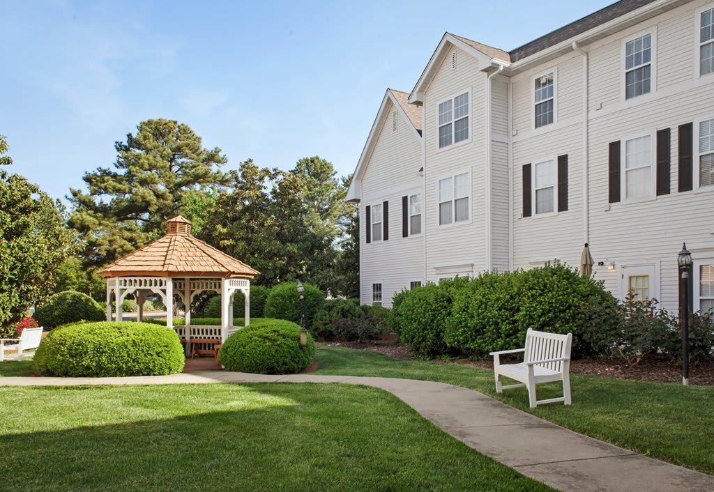 A serene garden area with a gazebo, pathway, and white bench next to a large building.