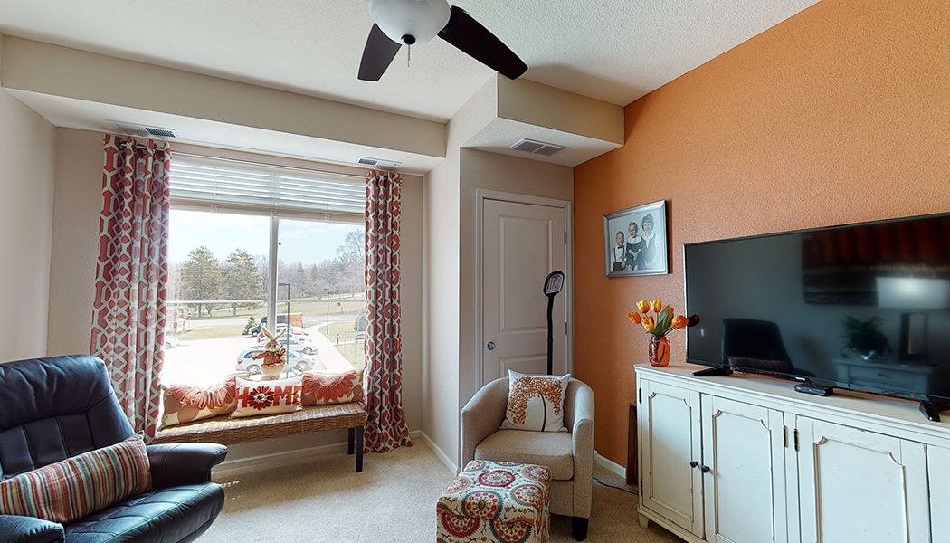 A cozy living room featuring a window seat, armchair, and television, with an orange accent wall and ceiling fan.