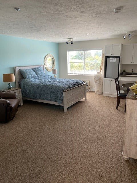 A spacious and organized bedroom with a bed, chair, lamp, and a large window allowing natural light.